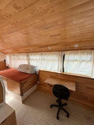 This image shows a cozy wooden room with a bed, a cushioned pillow, a small desk, a rolling chair, large windows, curtains, and patterned flooring.