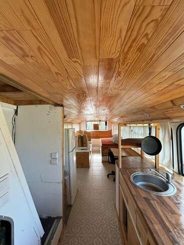 This image shows the interior of a converted bus or camper, featuring wood-paneled ceiling, compact kitchen, refrigerator, countertops, bed, desk, and light-colored patterned floor.