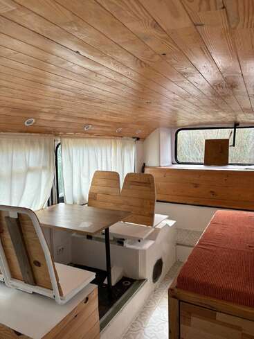 This cozy camper van interior features wooden ceiling and furniture, a small dining table with benches, a cushioned bench bed, large windows, and light curtains.