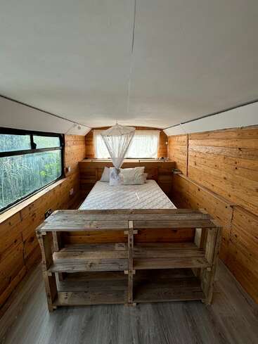 This cozy wooden room features a neatly made bed with white bedding, a small mosquito net, rustic shelves at the foot, and large windows overlooking greenery.