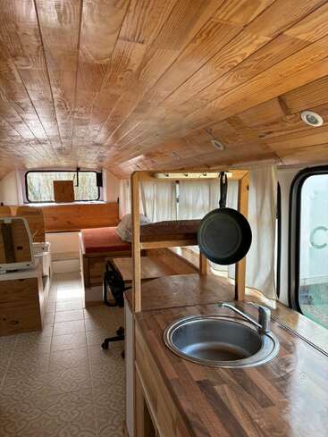 This image shows a cozy converted bus interior with wooden ceiling and counters, kitchenette with a sink, bed, windows with curtains, and a hanging frying pan.