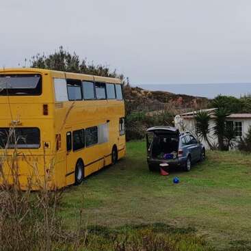 A imagem mostra um ônibus de dois andares estacionado em uma área gramada, com um carro por perto, cercado por uma pequena casa e árvores, em um céu nublado.