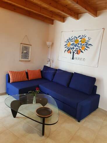 A cozy living room with a blue sofa, striped orange cushions, glass coffee table, wall art featuring oranges, wooden ceiling, and a single rose in a vase.