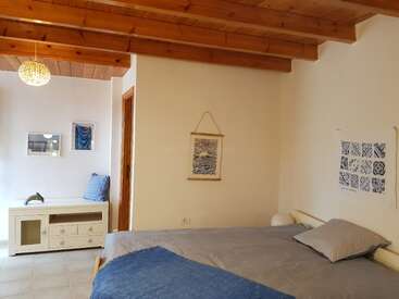This cozy bedroom features wooden ceiling beams, a comfortable bed with blue and gray bedding, simple wall art, and a small white bench with cushions.