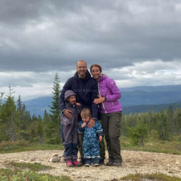 A family of four stands together outdoors, dressed in jackets and rain gear. Behind them are trees, mountains, and a cloudy sky, creating a serene atmosphere.