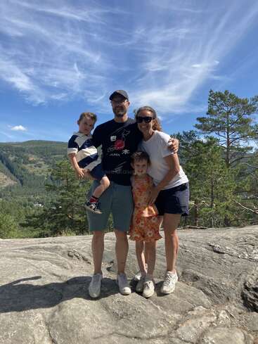 Une famille de quatre personnes souriantes pose au sommet d'une colline rocheuse sous un ciel bleu éclatant, entourée d'arbres verts et de montagnes, profitant d'une aventure en plein air sous le soleil.