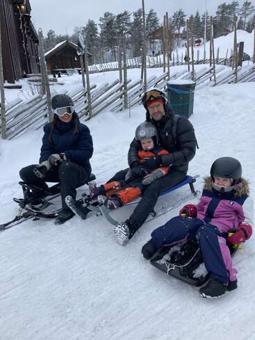 Une famille de quatre personnes, vêtues de tenues d'hiver chaudes et de casques, est assise sur des luges dans un paysage enneigé, souriante et prête à s'amuser.