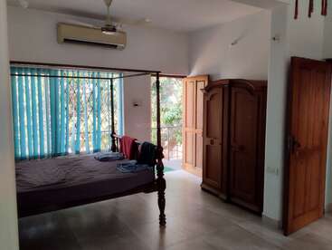 This image shows a simple bedroom with a wooden bed, blue vertical blinds, wooden wardrobe, ceiling fan, air conditioner, and an open door with sunlight coming in.