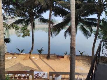 Tall palm trees line a sandy area by a calm river. The tranquil water reflects greenery, while scattered debris and a stairway complete the serene scene.
