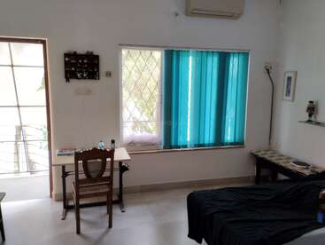 Essa é uma sala de estar bem iluminada e arrumada, com uma mesa de estudo, cadeira de madeira, persianas verticais azuis, sofá preto, prateleiras de parede e um ar condicionado split.