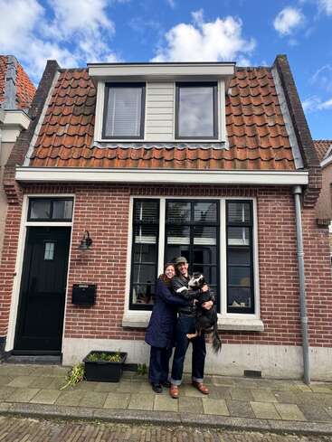 A smiling couple stands in front of a charming brick house with a red tiled roof, holding a black and white dog under a bright blue sky.