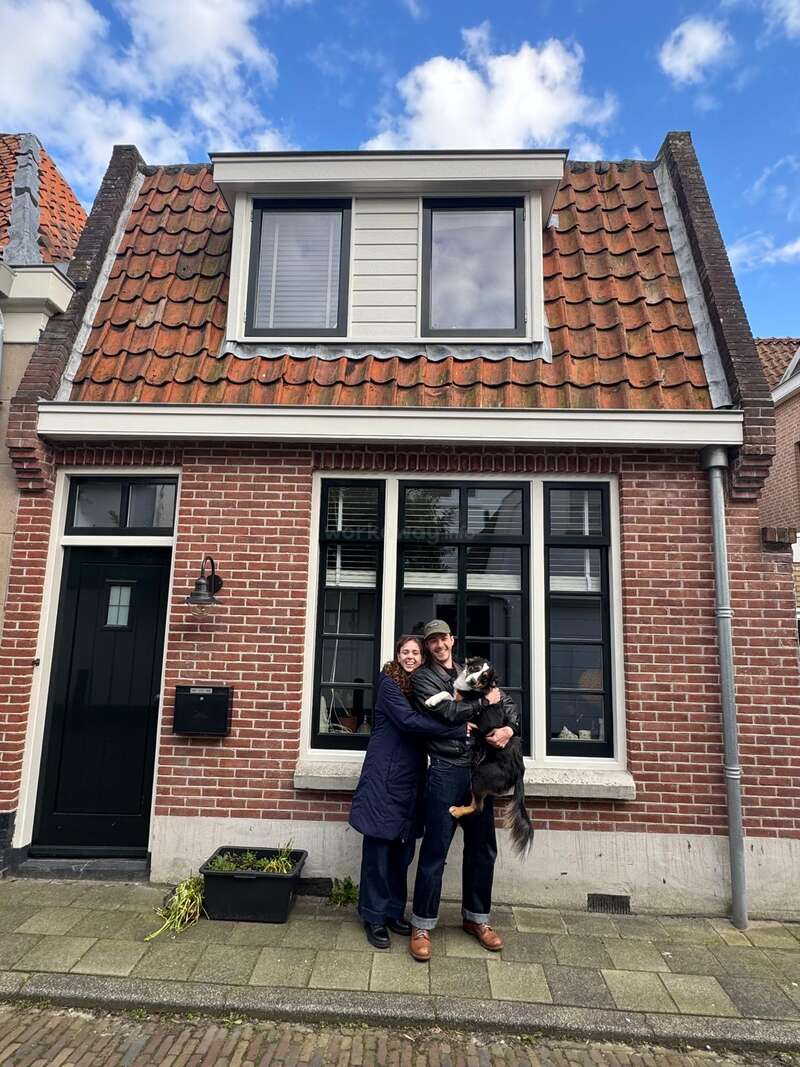 A smiling couple stands in front of a charming brick house with a red tiled roof, holding a black and white dog under a bright blue sky.