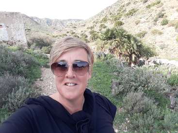 A imagem mostra uma mulher tirando uma selfie em frente a uma paisagem desértica com vegetação, usando óculos escuros e um capuz preto.
