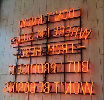 A imagem mostra um letreiro de neon com uma citação em uma parede de madeira, o texto está de cabeça para baixo e diz: "NÃO SEI PARA ONDE VOU IR A PARTIR DE AQUI, MAS PROMETO QUE NÃO VAI SER CHATO".