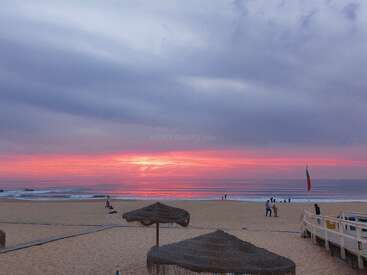 A serene beach at sunset, sky painted with pink and purple hues. People stroll on the sand, enjoying the calm atmosphere and gentle ocean waves.