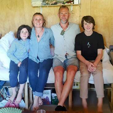 Une famille de quatre personnes est assise en rangs serrés sur un lit dans une chambre confortable en bois, et sourit chaleureusement. Des étagères contenant des vêtements pliés sont visibles au-dessus d'eux.