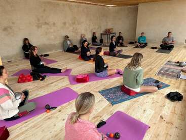 Eine Gruppe von Menschen sitzt auf Yogamatten in einem ruhigen Raum mit Holzboden und nimmt an einer Meditations- oder Klangheilungssitzung teil, jeder hält eine Metallschale in der Hand.