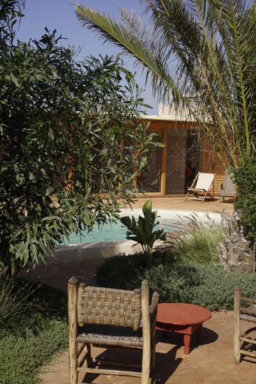 Eine gemütliche Terrasse mit rustikalen Holzstühlen und einem roten Tisch bietet einen Blick auf einen kleinen Pool, der von üppigen grünen Pflanzen, Palmen und einem Holzhaus umgeben ist.