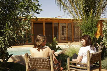 Zwei Frauen sitzen auf geflochtenen Stühlen, schauen sich an und lächeln, umgeben von üppigem Grün, mit einem Swimmingpool und einem Holzhaus im Hintergrund.