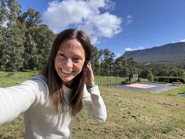 A woman smiles outdoors, taking a selfie on a sunny day. Behind her is a basketball court, green trees, grassy field, and distant blue mountains.