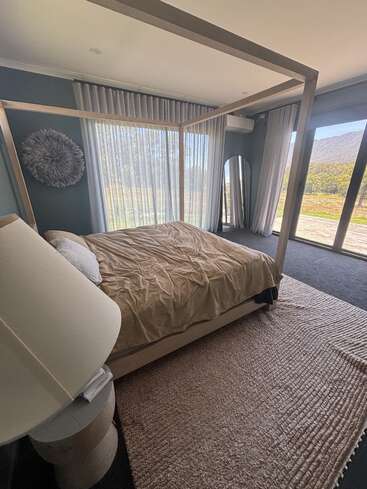 A modern bedroom with a four-poster bed, tan bedding, soft rugs, floor-length curtains, a large window with scenic views, and a decorative wall piece.