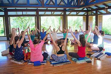 Um grupo de pessoas senta-se em círculo em uma sala iluminada e aberta. Elas dão as mãos, levantam os braços e parecem estar participando de uma alegre atividade em grupo.