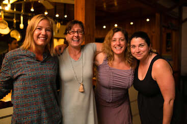 Quatro mulheres estão juntas em um ambiente interno, sorrindo e rindo. A iluminação quente e a atmosfera aconchegante sugerem uma reunião amigável, possivelmente em uma cozinha ou área de jantar.