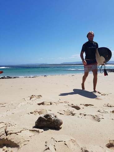 The image depicts a man walking on a sandy beach, carrying a surfboard under his arm, with a serene ocean view and a clear blue sky in the background.