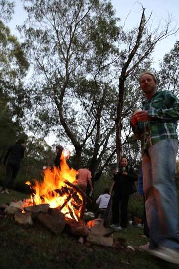 The image depicts a group of people gathered around a campfire in a wooded area, with trees visible in the background.