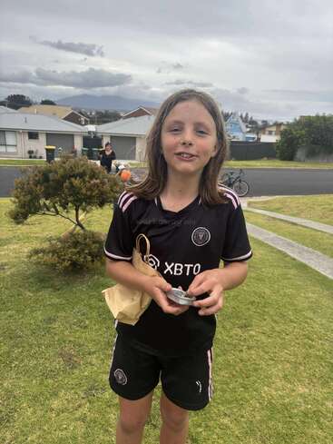 A young child with long hair stands on a lawn, wearing a black sports uniform. They hold a small item and brown paper bag, smiling outdoors.
