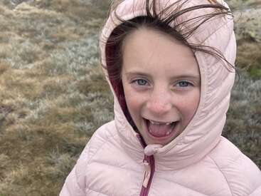 A joyful child in a pink puffer jacket with a hood smiles brightly outdoors. Her hair is windswept, and the background is grassy and natural.