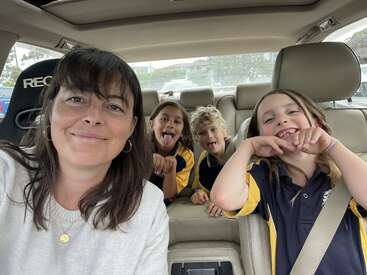 A woman and three children sit in a car. The kids wear uniforms and make playful faces, while the woman smiles, taking a cheerful selfie together.