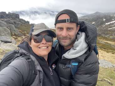A smiling couple takes a selfie while hiking in a rocky, mountainous landscape. They wear hats, sunglasses, and jackets, with cloudy skies and misty mountains behind.