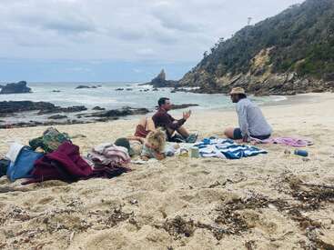 Eine Gruppe von Leuten entspannt sich an einem Sandstrand, umgeben von Handtüchern, Taschen und Bechern. Sie unterhalten sich und genießen die felsige Küste unter bewölktem Himmel.
