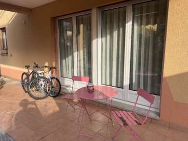 Un patio soleado cuenta con una pequeña mesa redonda rosa y dos sillas a juego. Dos bicicletas están aparcadas cerca, contra una pared con grandes puertas de cristal.