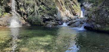 Ein klarer, ruhiger Pool liegt unter Wasserfällen, umgeben von felsigen Klippen und üppigem Grün. Das Sonnenlicht spiegelt sich auf dem Wasser, während zwei Leute die Felsen erkunden.
