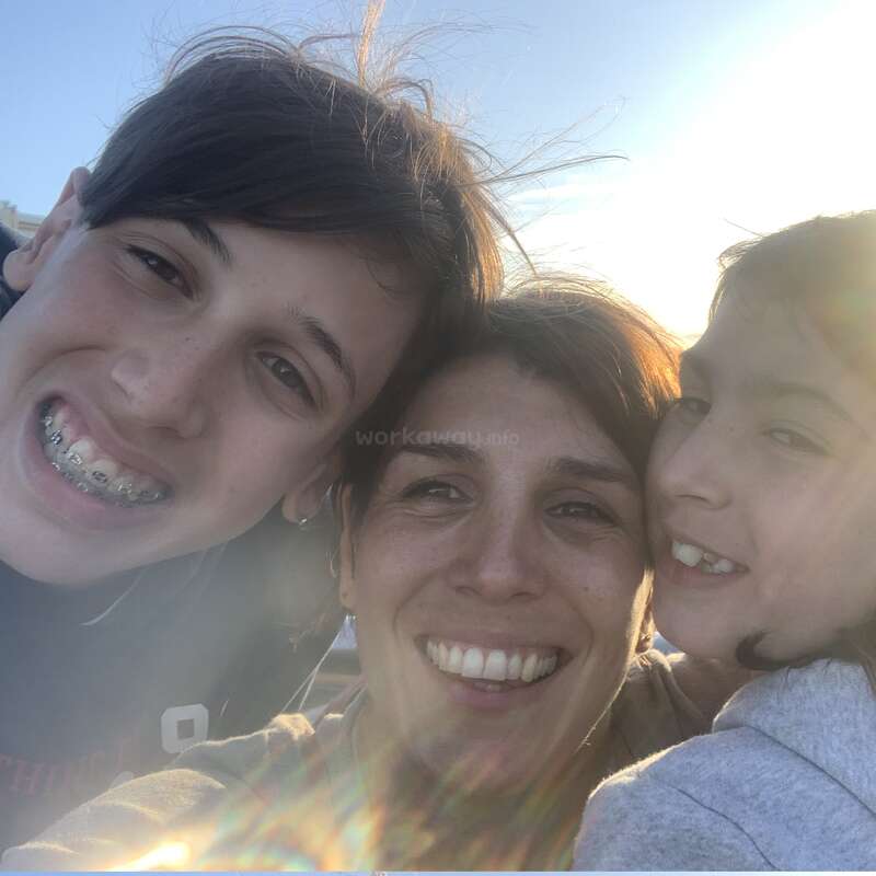 Três pessoas, duas crianças e um adulto, sorriem alegremente juntas ao ar livre. A luz do sol cria um brilho quente e um reflexo na lente, realçando o momento feliz e íntimo que vivem.
