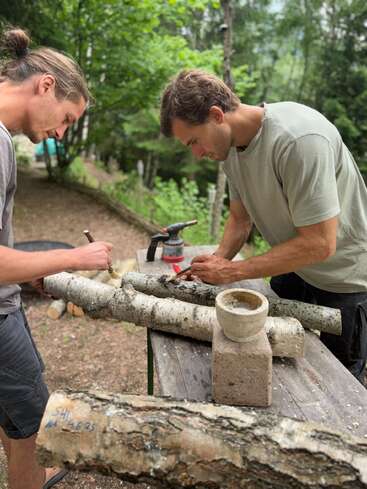 Two men work intently outdoors, drilling or carving into birch logs on a wooden table, surrounded by lush green trees, with tools and a mortar present.