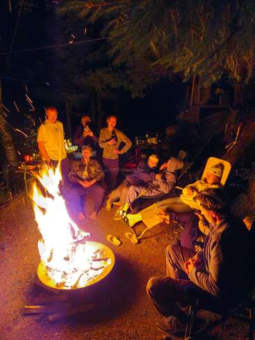 A group of friends gather around a glowing campfire at night, laughing, talking, and relaxing under trees, enjoying warmth, light, and each other's company outdoors.