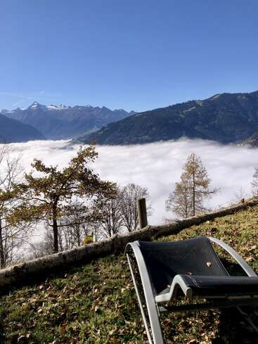 A lounge chair rests on grassy ground, overlooking stunning mountains covered in snow, fog-filled valley, bare trees, and a clear blue sky, creating a serene scene.