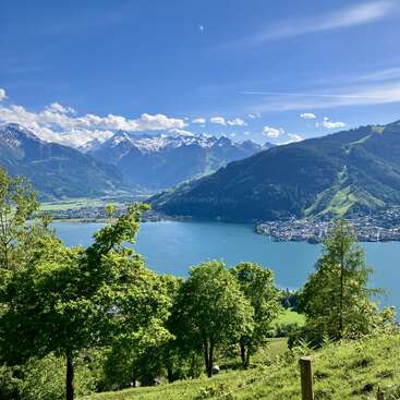 A stunning landscape features a pristine blue lake, surrounded by lush green hills, vibrant forests, and majestic snow-capped mountains beneath a bright, clear sky.