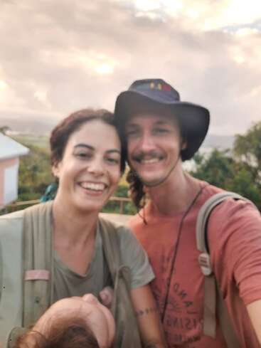 A smiling couple takes a selfie outdoors at sunset. The woman carries a baby in a front carrier. They look happy, enjoying a beautiful, natural setting together.