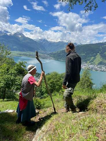 Una anciana con un bastón y un hombre más joven se encuentran en una ladera cubierta de hierba, contemplando un impresionante lago y un paisaje de montañas nevadas bajo un cielo azul.