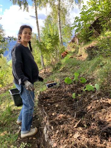 Una mujer con trenzas, guantes y un jersey oscuro está trabajando en el jardín en una ladera rodeada de árboles verdes, bajo un cielo despejado, y sonríe a la cámara.