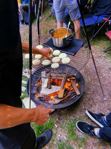 Un grupo de personas está al aire libre, tostando pan sobre una hoguera. Una olla con guiso o sopa reposa sobre una parrilla, creando un ambiente acogedor de acampada.