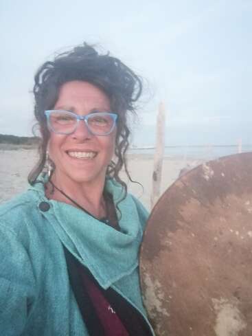 Uma mulher sorridente, com cabelos cacheados e óculos azuis, está em uma praia, segurando um grande tambor redondo. Ela veste uma roupa azul-petróleo e parece feliz ao ar livre.