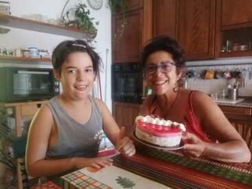 Uma mulher e uma criança sorriem sentadas à mesa numa cozinha aconchegante. A mulher segura um bolo decorado. A criança levanta o polegar. Um momento feliz.