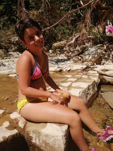 Uma jovem de biquíni colorido está sentada em pedras à beira de um riacho raso, sorrindo, cercada pela natureza, pela luz do sol, pelas rochas e pelas árvores verdes ao fundo.