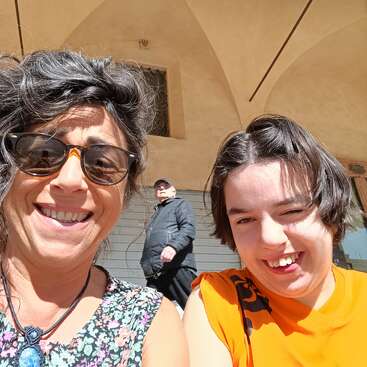 Eine Frau mit Sonnenbrille und einem geblümten Oberteil lächelt neben einem jungen Mann in einem orangefarbenen Hemd; beide genießen die Sonne, im Hintergrund steht ein älterer Mann.