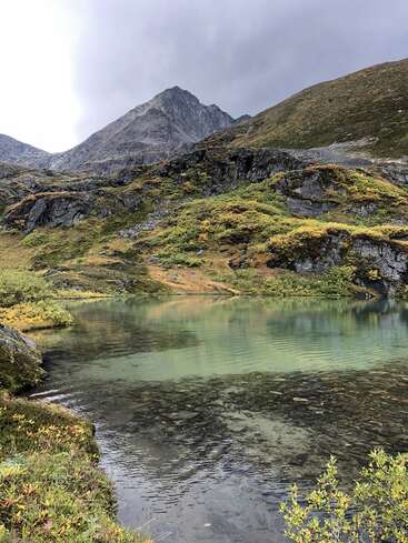L'image représente un paysage montagneux serein, avec un lac entouré d'une végétation luxuriante et de rochers escarpés, le tout dans un ciel couvert.